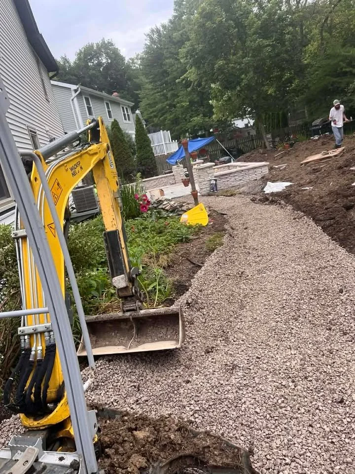 A construction site with a small yellow excavator working on a gravel pathway in a backyard, with construction materials and workers present, surrounded by green trees and residential houses.