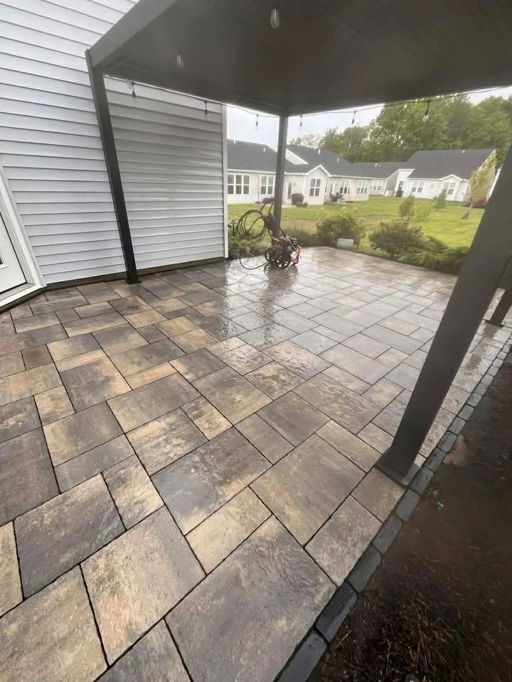 A wet patio with beige and gray stone pavers, a small child's tricycle, and a garden hose, under a black patio cover, with a grassy backyard and white houses in the background.
