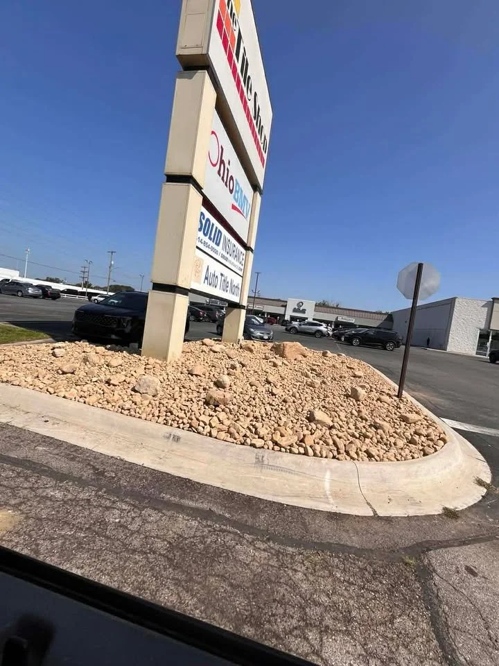 A parking lot with a large roadside sign advertising real estate companies and auto insurance. The sign is mounted on a concrete base surrounded by a bed of rocks. There are many parked cars and a building in the background under a clear blue sky.