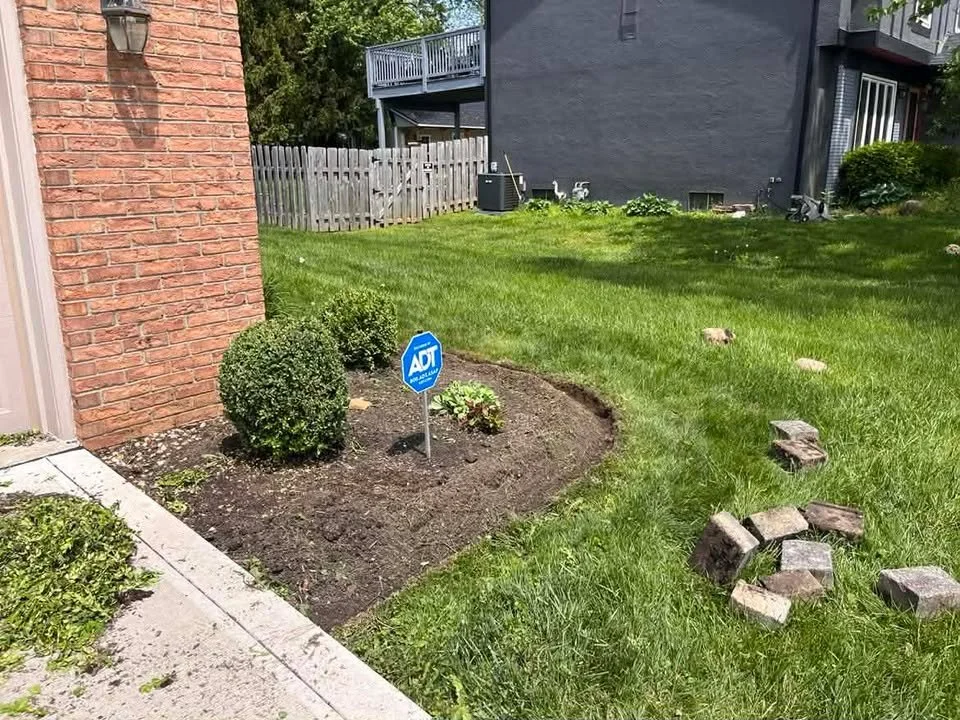 A small garden bed with two neatly trimmed bushes, a small flowering plant, and a security sign near a brick building, with a grassy yard extending to a fence and neighboring house in the background.