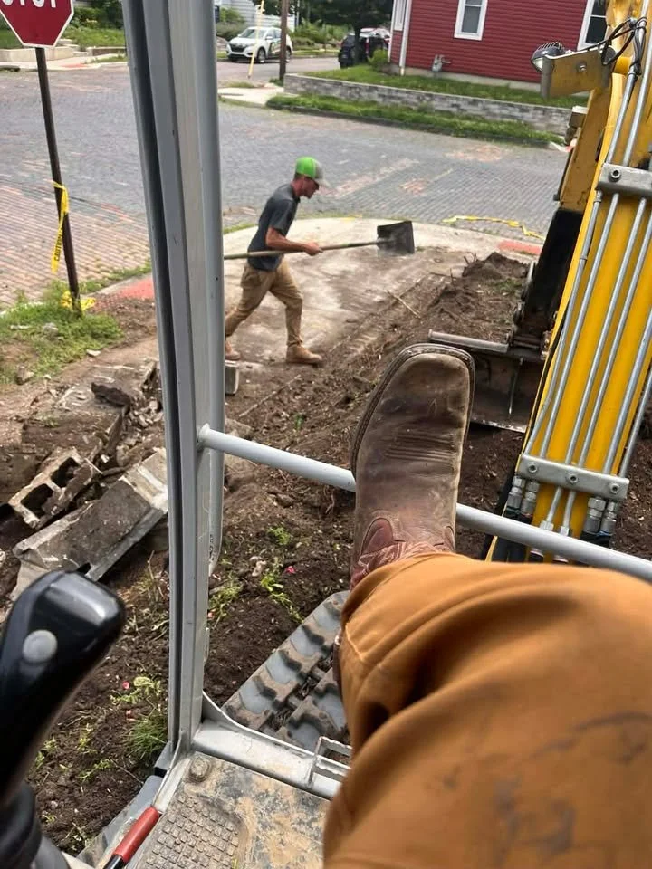 A construction worker in a gray shirt and wearing a green hard hat is shoveling dirt at a construction site near a red building. The perspective is from inside a small excavator, showing the operator's foot resting on the machine's control panel.
