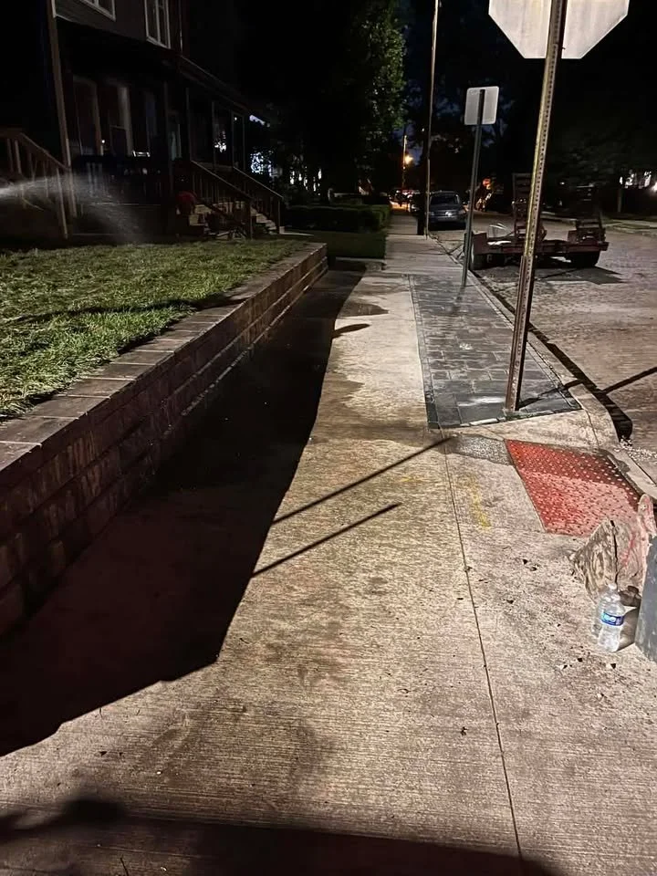 Nighttime view of a sidewalk beside a street with parked cars, streetlights illuminating the scene, and a partially covered sidewalk with a red textured ground surface near a bent street sign. A water bottle is on the sidewalk near the curb.