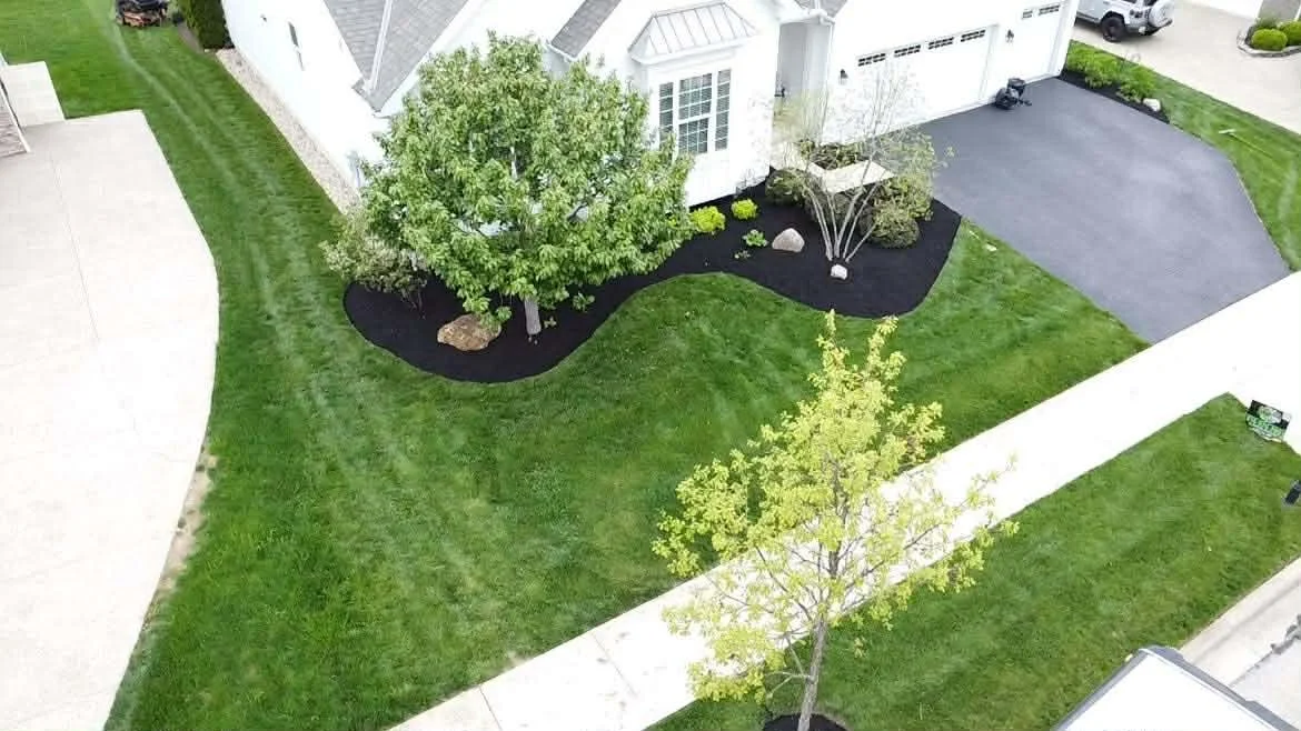 An aerial view of a backyard with a grassy lawn, landscaped garden beds with small trees and rocks, a concrete walkway, and a driveway with a parked truck.