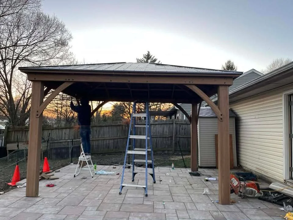 A person working on constructing a wooden gazebo on a patio during sunset, with tools and equipment scattered around.