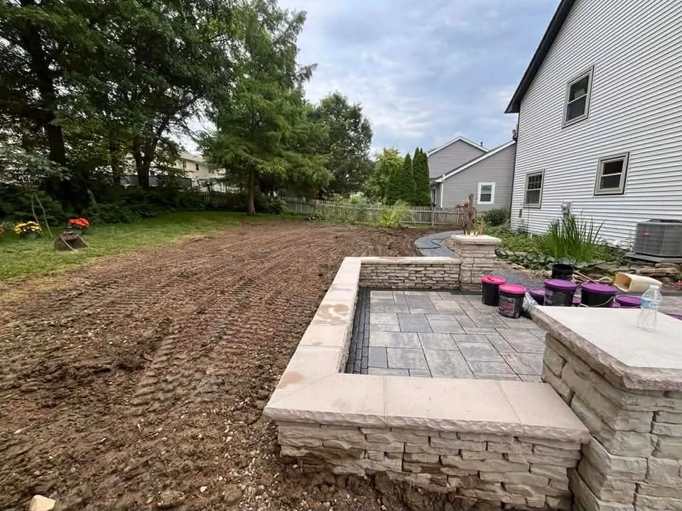 Backyard under renovation with soil prepared for landscaping, a paver stone patio area, and construction materials like buckets and a water bottle nearby.