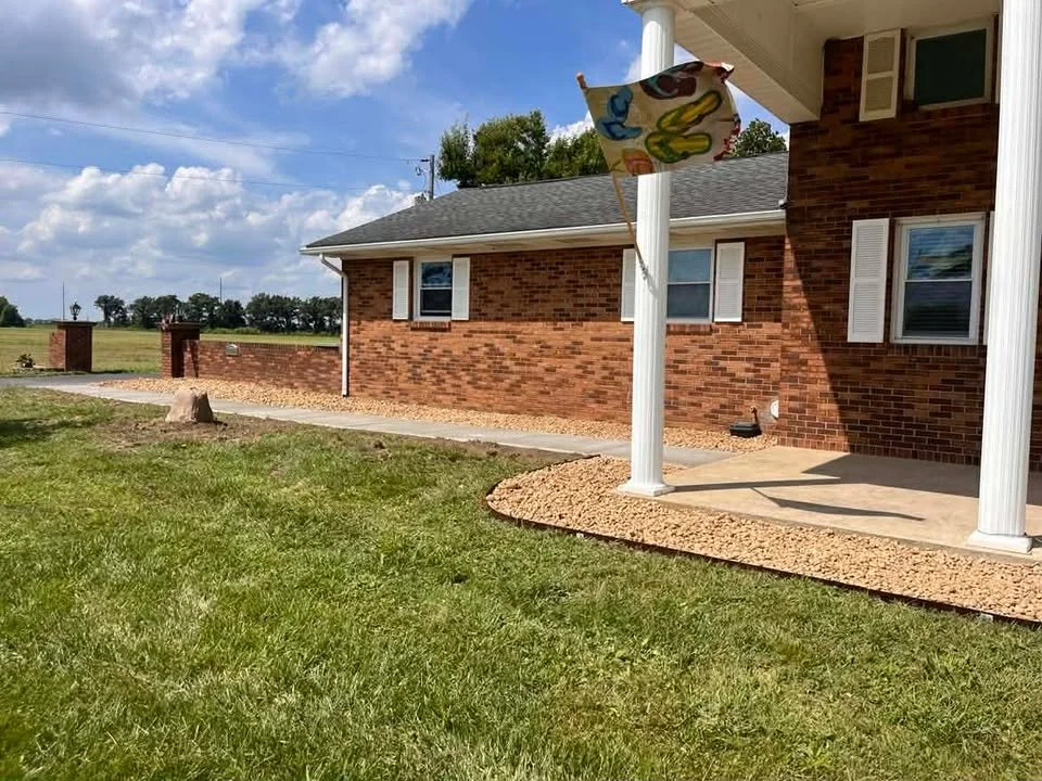 A brick house with a porch, white columns, and a decorative flag hanging from one of the columns. The house is surrounded by a grassy yard and a gravel pathway. The sky is partly cloudy.