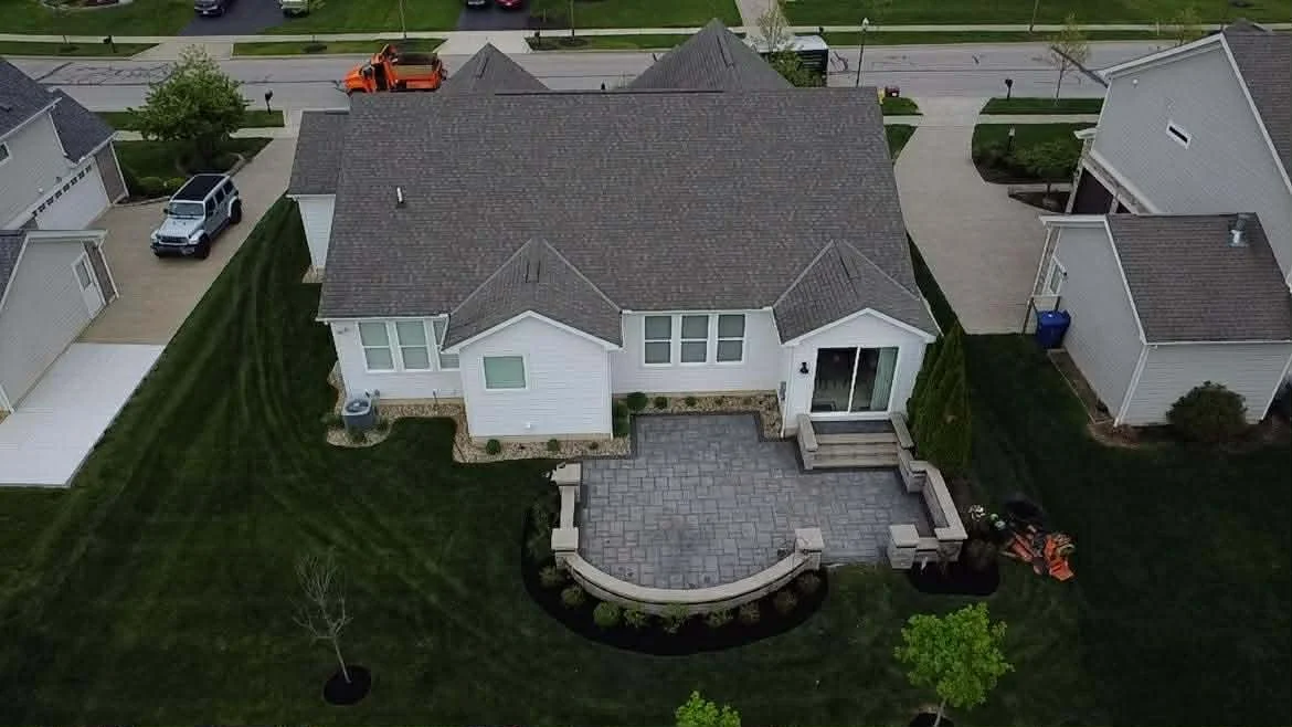 Aerial view of a suburban house with a newly paved backyard patio, green lawn, and neighboring houses in the background.