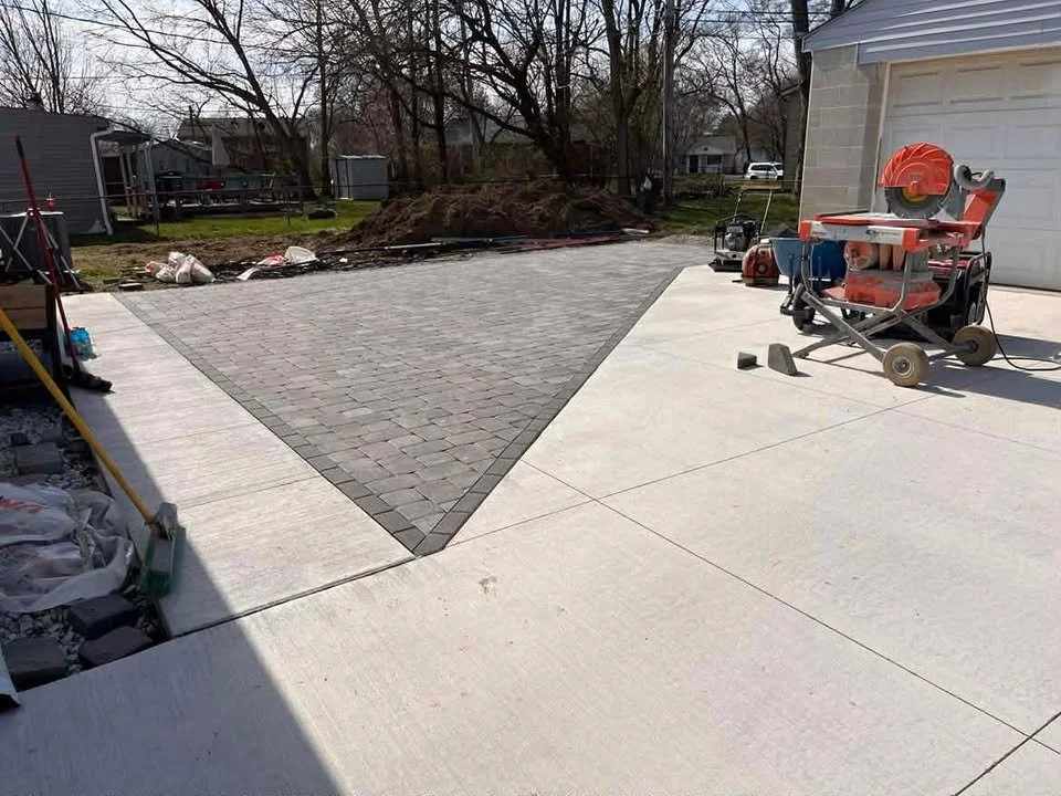 A backyard scene showing a newly paved driveway with a section of brick pavers and a concrete section, construction tools, a worktable with saw, and piles of materials, with some trees and houses in the background.