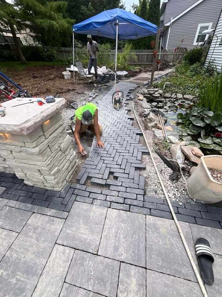 Person laying down black bricks to create a patterned pathway in a backyard garden, with a pond containing lily pads on the right, and another person working under a blue canopy in the background.