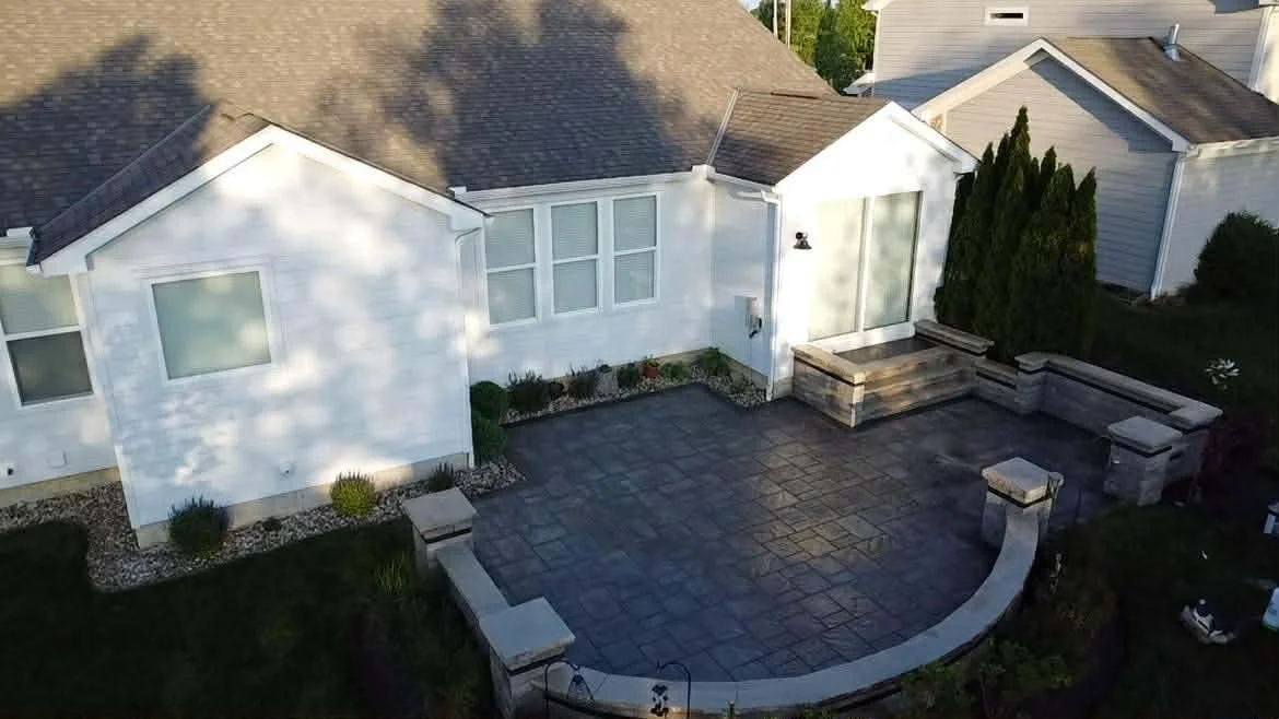 A backyard patio with a dark stone surface, surrounded by a curved stone bench and stairs leading to a white house with multiple windows and a glass sliding door.