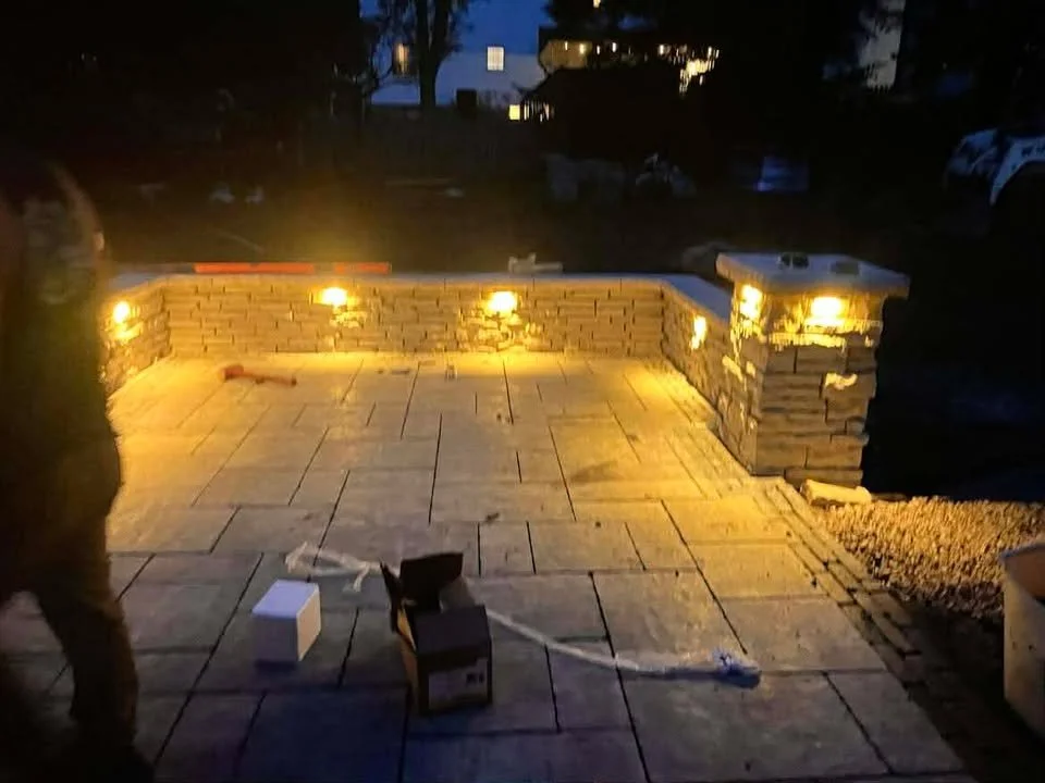 A newly built outdoor patio with stone brick walls and seating, illuminated by warm yellow lighting, at dusk or night with some construction materials and tools visible.