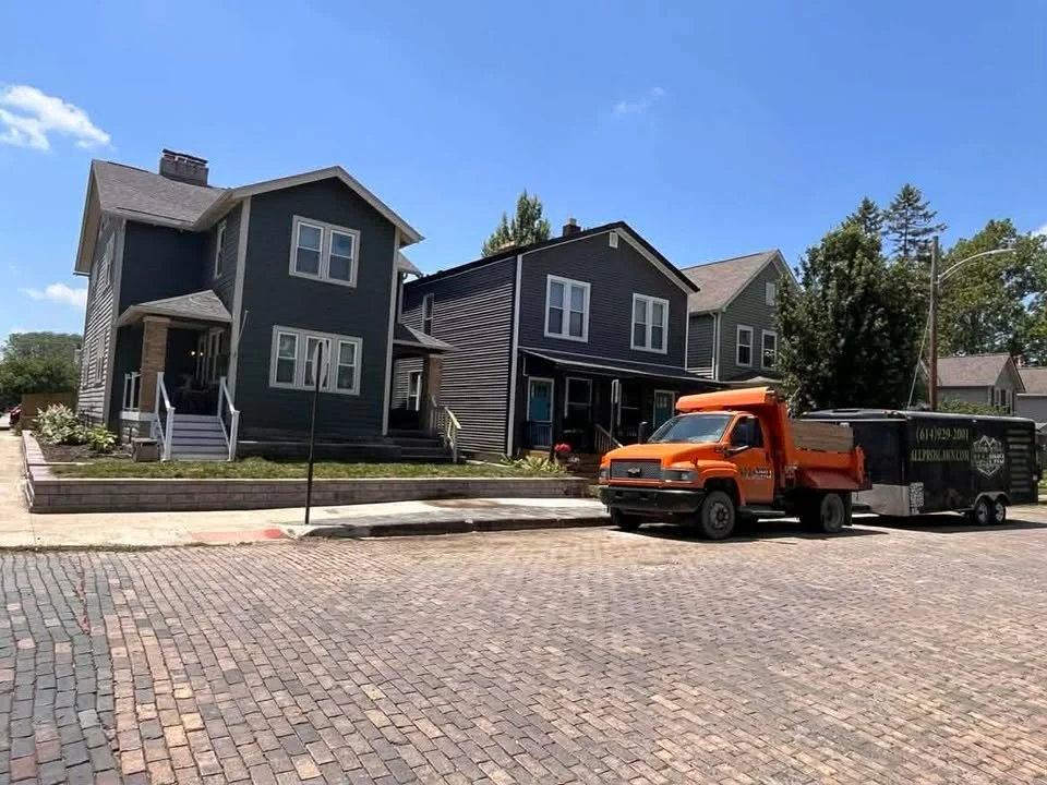 Two modern houses with front steps and small front yards, parked orange truck and black trailer on brick-paved street under a blue sky.