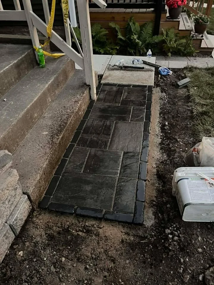 Newly laid black paver stones on a walkway next to steps, with construction materials and tools nearby, including a green bottle, water bottles, and a white container.