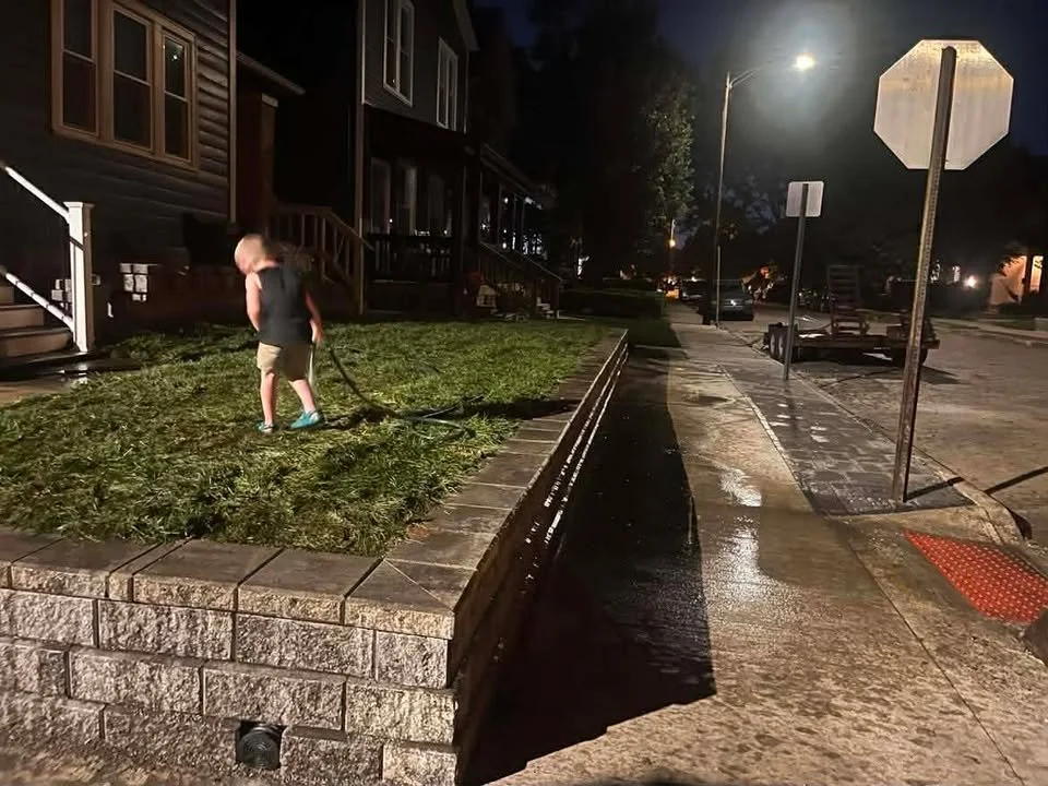 A young boy wearing a black shirt and beige shorts is watering or cleaning a small grassy area with a hose on a residential street at night. Houses and streetlights line the street, which is wet from recent watering or cleaning.