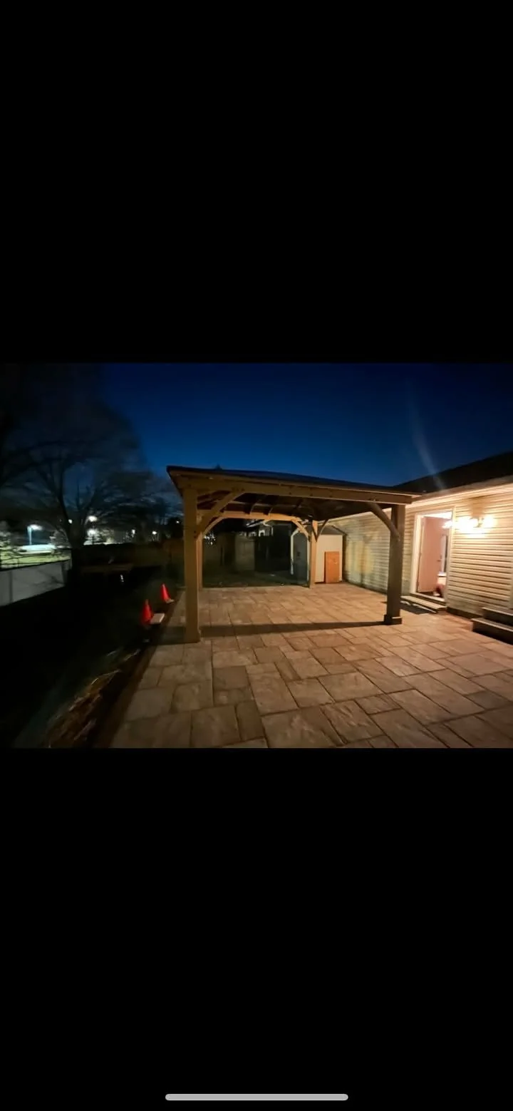 Empty backyard patio with a wooden pergola, stone flooring, and house siding illuminated by a porch light, at dusk.
