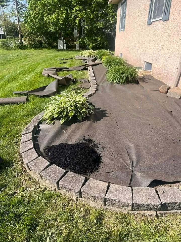 A garden bed with black landscape fabric partially peeled back revealing a patch of black compost or soil. The bed is bordered by gray bricks and contains green plants. The surrounding area has green grass and leafy trees.