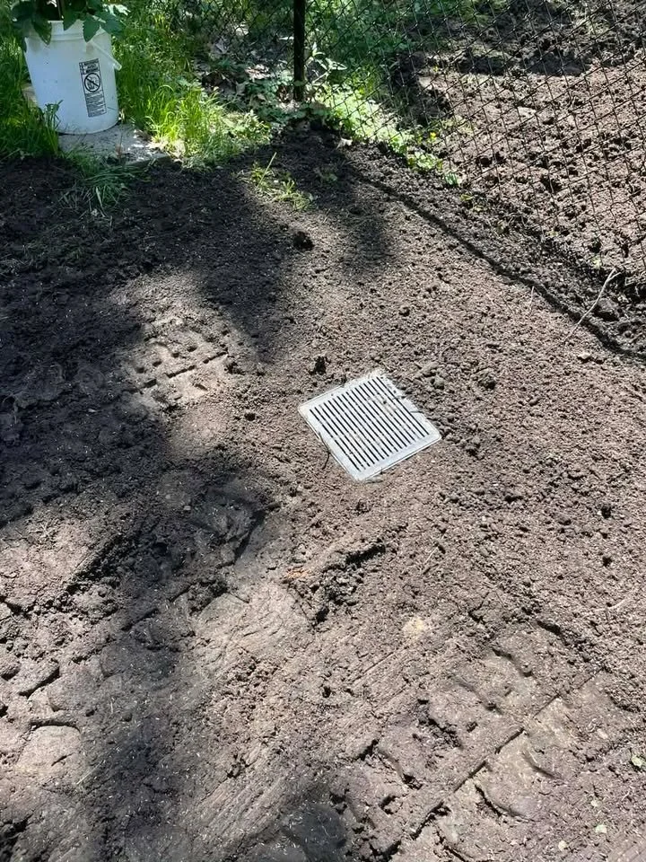 Freshly tilled soil with a white drainage grate in the center, fenced yard with grass and gardening supplies in the background.