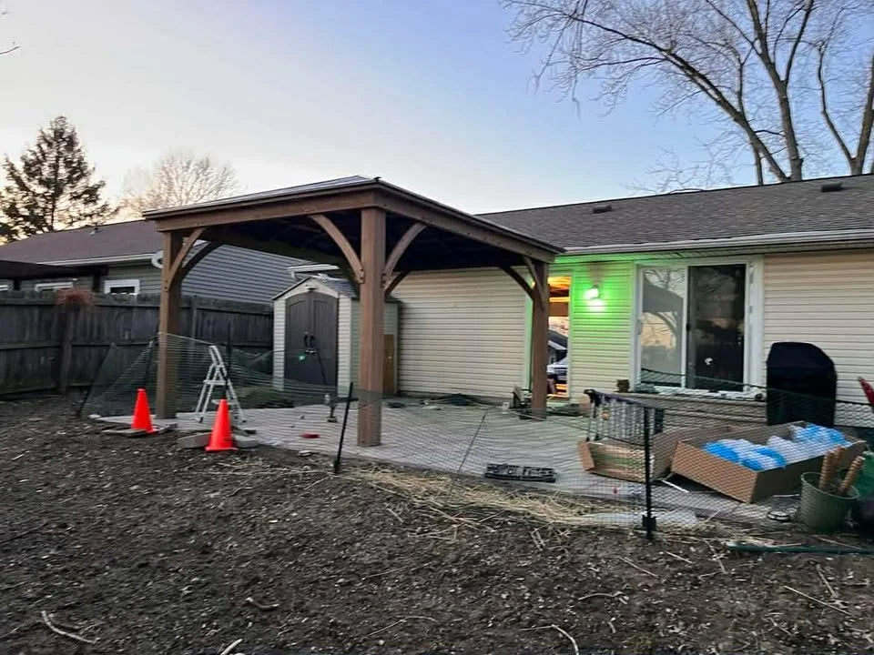 Backyard patio under construction with a wooden pergola, orange traffic cones, gardening tools, and a fenced-off area in front of a house with sliding glass doors.