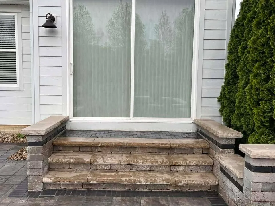 Front porch with stone steps leading to a sliding glass door, with a black outdoor light fixture on the left and tall green shrubbery on the right.