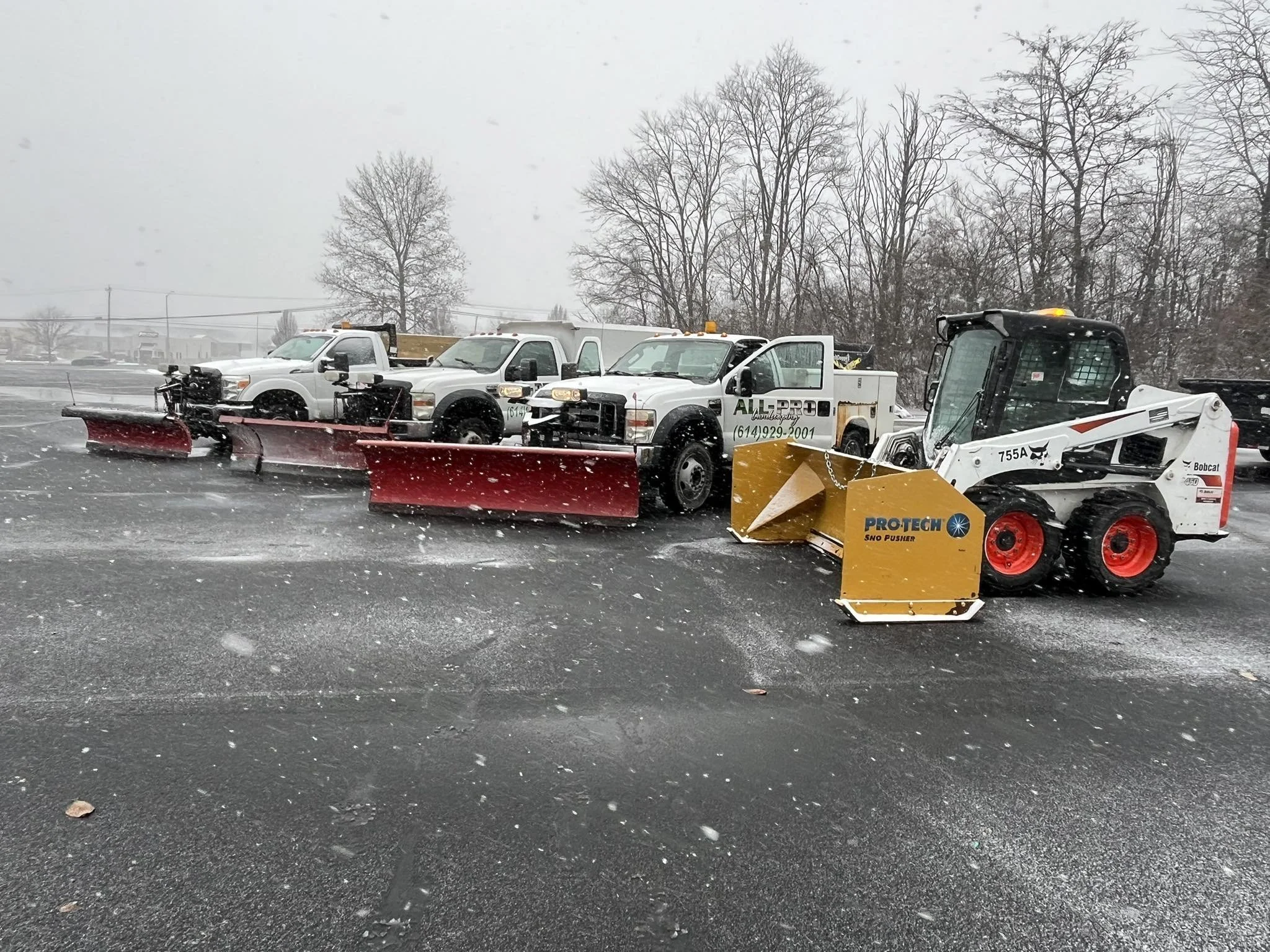 Snow-covered parking lot with three pickup trucks equipped with red snow plows and a Bobcat skid-steer loader with a yellow snow pusher attached, parked in a row, with leafless trees in the background under a gray, snowy sky.
