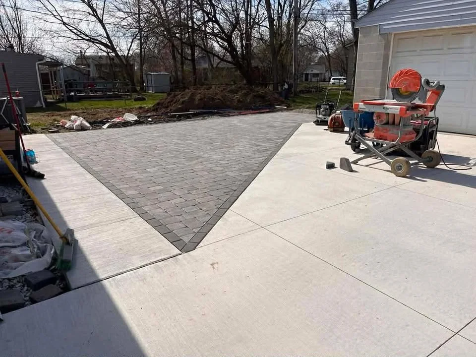 Newly paved driveway with a section of gray brick pavers and surrounding concrete, construction tools and materials present, outdoors in a residential backyard with trees and neighboring houses.