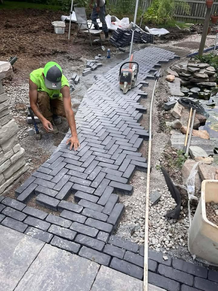 A worker installing a herringbone pattern brick walkway in a backyard garden, with tools and materials visible around him.