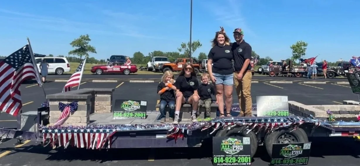 Group of five people, including children and adults, standing and sitting on a float decorated with American flags and patriotic colors at a parade. Many cars and spectators in the background on a clear, sunny day.