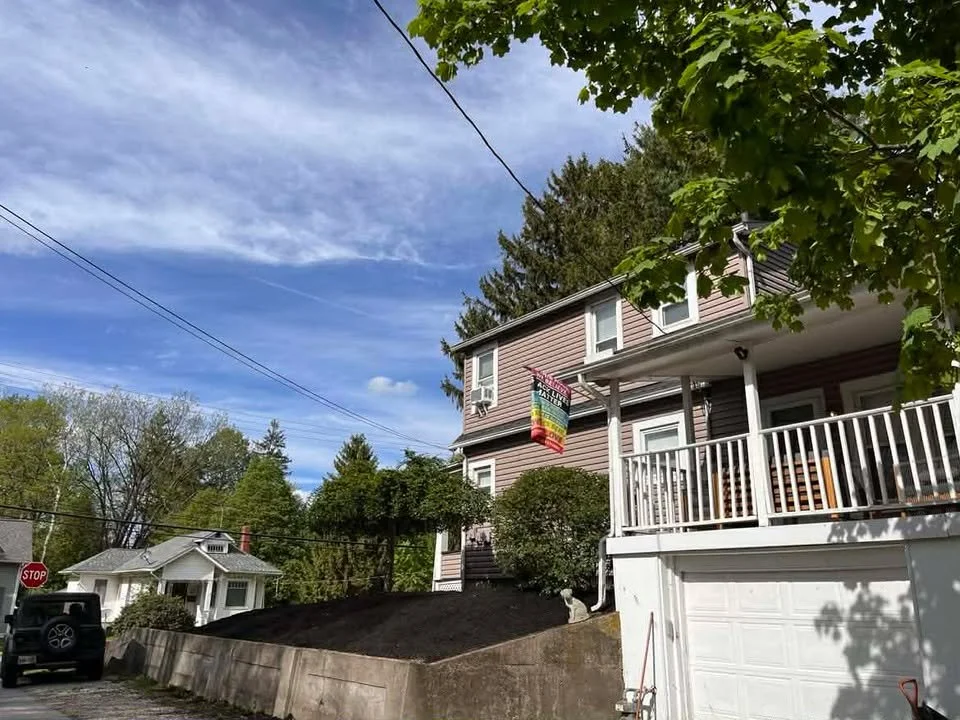 A house with a colorful rainbow flag hanging from the porch, surrounded by greenery, under a partly cloudy sky.