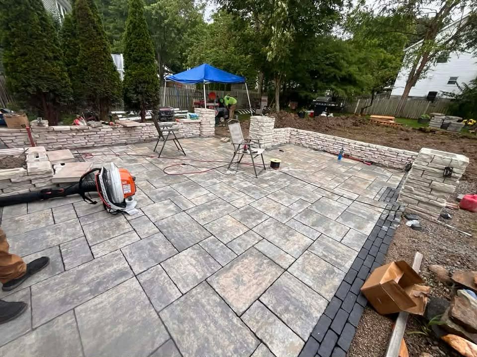 Under construction backyard patio with stone walls and paving stones, construction tools, and workers installing the area.