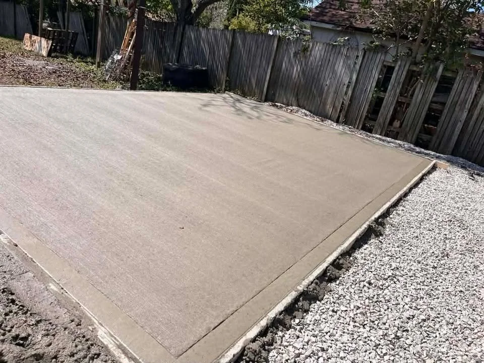 Freshly poured concrete slab with gravel surrounding it, in a backyard or garden area, with a wooden fence and trees in the background.