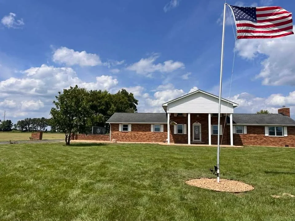 A large American flag flying on a flagpole in front of a brick house with an open lawn and blue sky with some clouds.