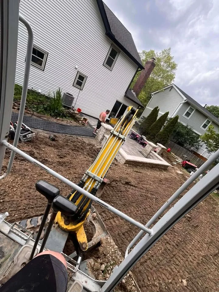 View from construction equipment showing a backyard with a patio, some newly planted bushes, and a person working near the house. There are construction tools and dirt in the foreground.
