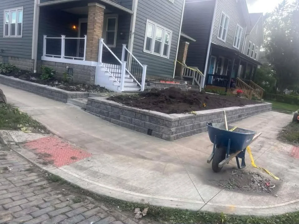 Construction site with newly built brick retaining wall, dirt area, wheelbarrow, and house with porch and stairs