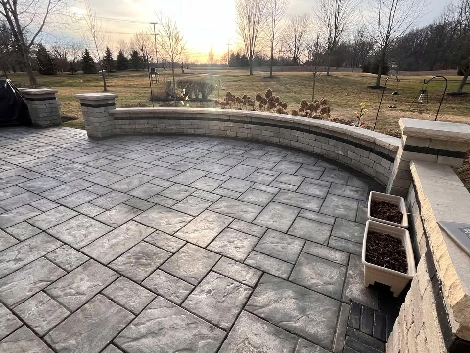 A stone patio with a curved brick wall, several trees and an open grassy area in the background at sunset.
