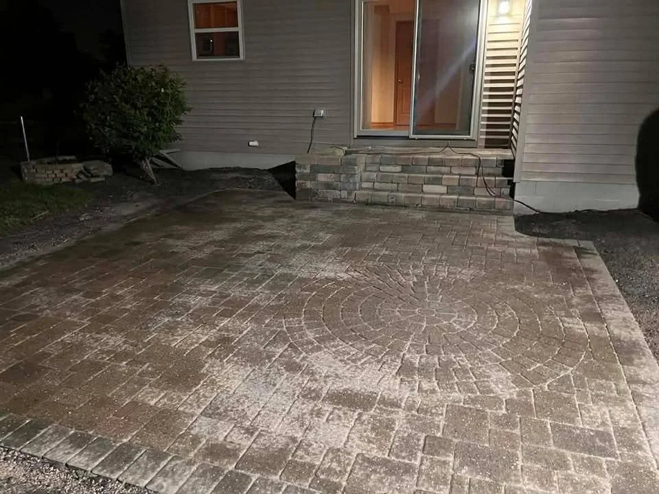 Nighttime view of a newly installed brick patio in front of a house with sliding glass door, a small set of brick steps, and a bush on the left side.