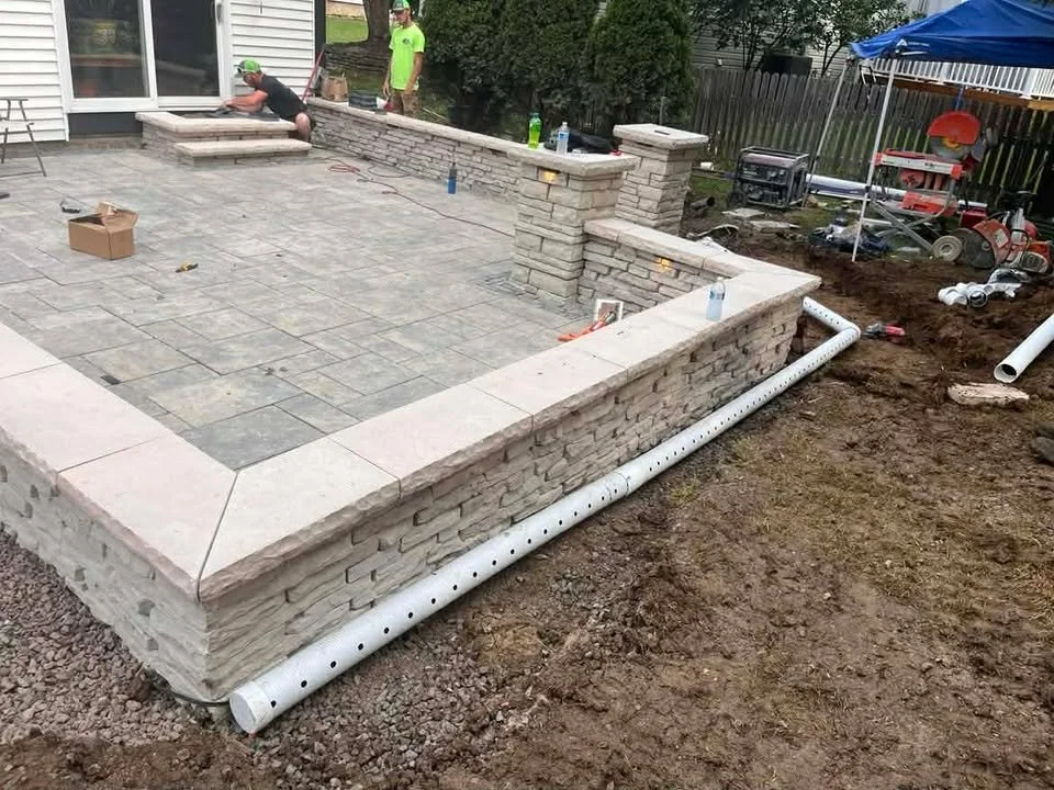 Workers installing stone paving on a backyard patio, with a stone wall and drainage pipe visible.