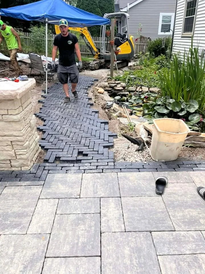 People installing an outdoor stone patio with a herringbone pattern, next to a garden pond and a small excavator in the yard.