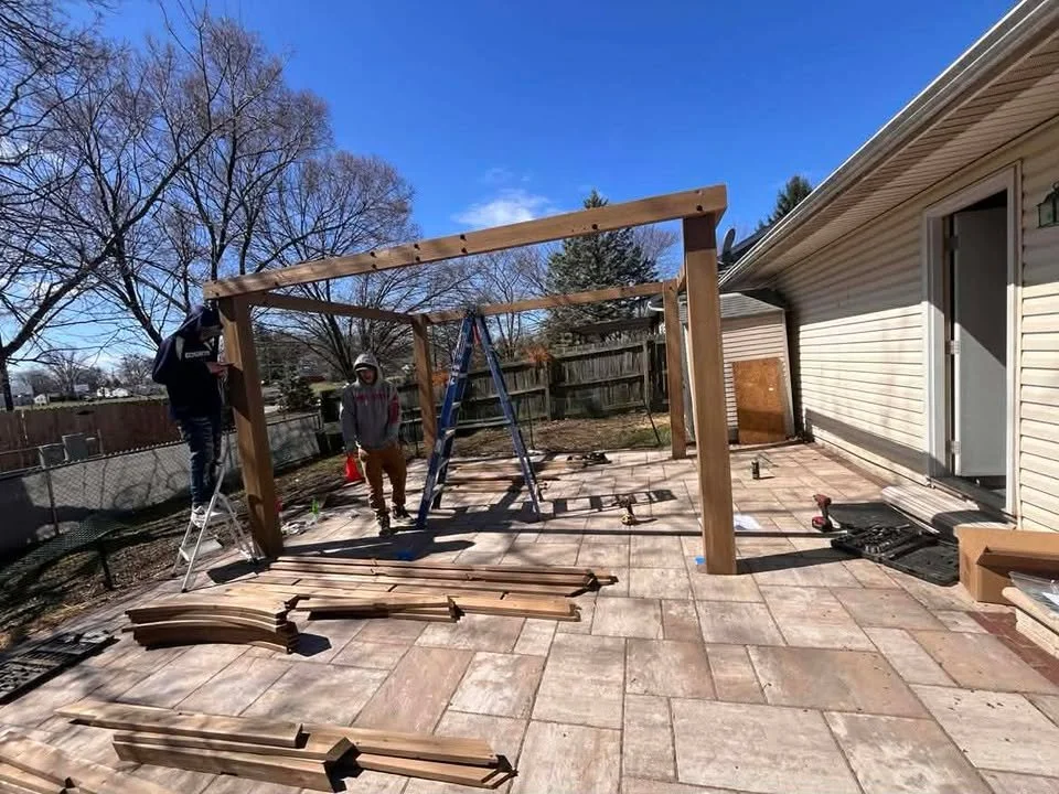 Three men building a wooden structure outdoors on a patio with pavers, with a ladder, tools, and wood scattered around.