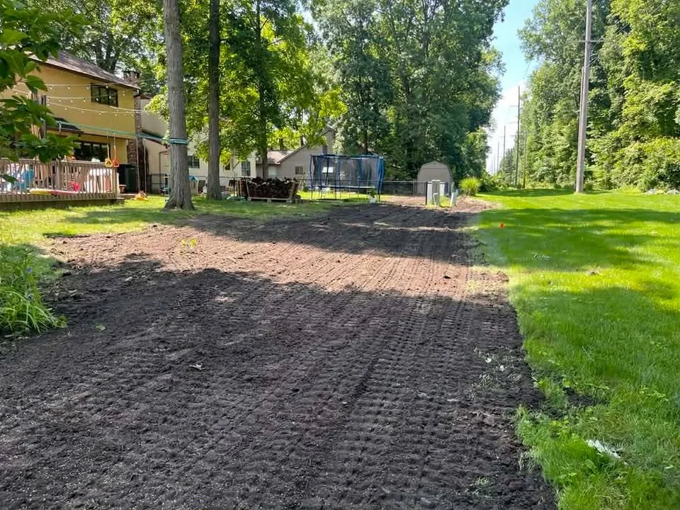 Backyard with dirt covered area, green grass, trees, a trampoline, a basketball hoop, and a house with a deck.