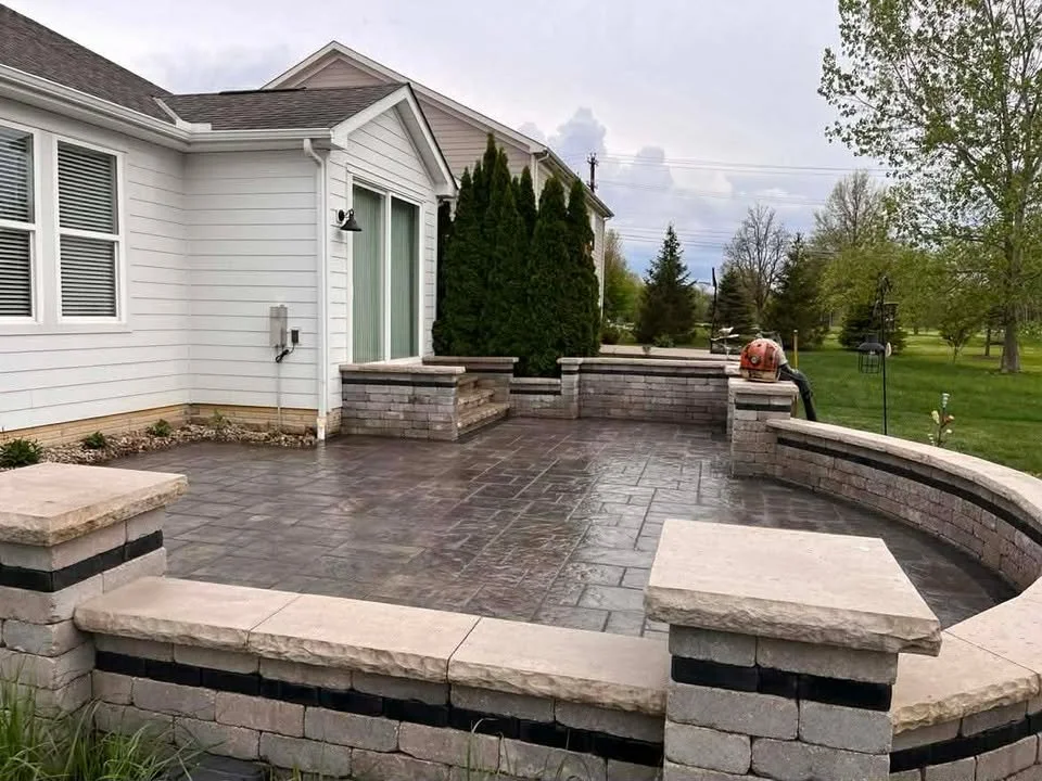 Backyard patio with dark stamped concrete, brick border wall, and white house exterior with sliding glass door.