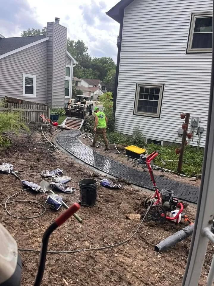 Construction workers are installing a new brick pathway in a backyard. They are spreading and leveling the dark bricks that curve around the yard. Construction tools, buckets, and materials are scattered around the area, with a small excavator in the