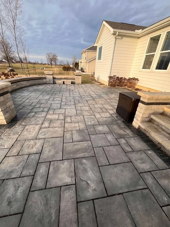 Newly paved backyard patio with gray stone tiles, brick walls, and steps leading up to the house, with a lawn and trees in the background.