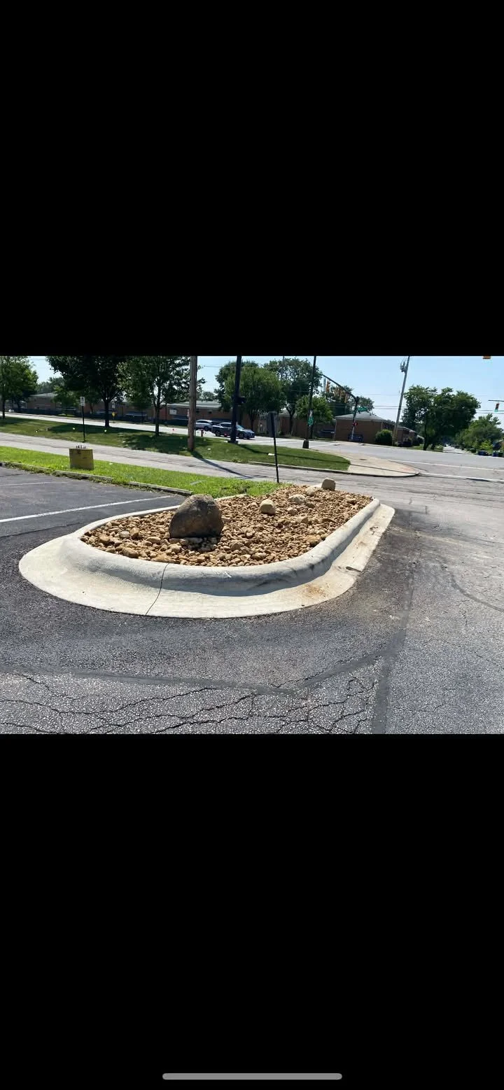 Empty parking spot with a small landscaped island filled with rocks and three large rocks, in a parking lot near a street with trees and buildings in the background.