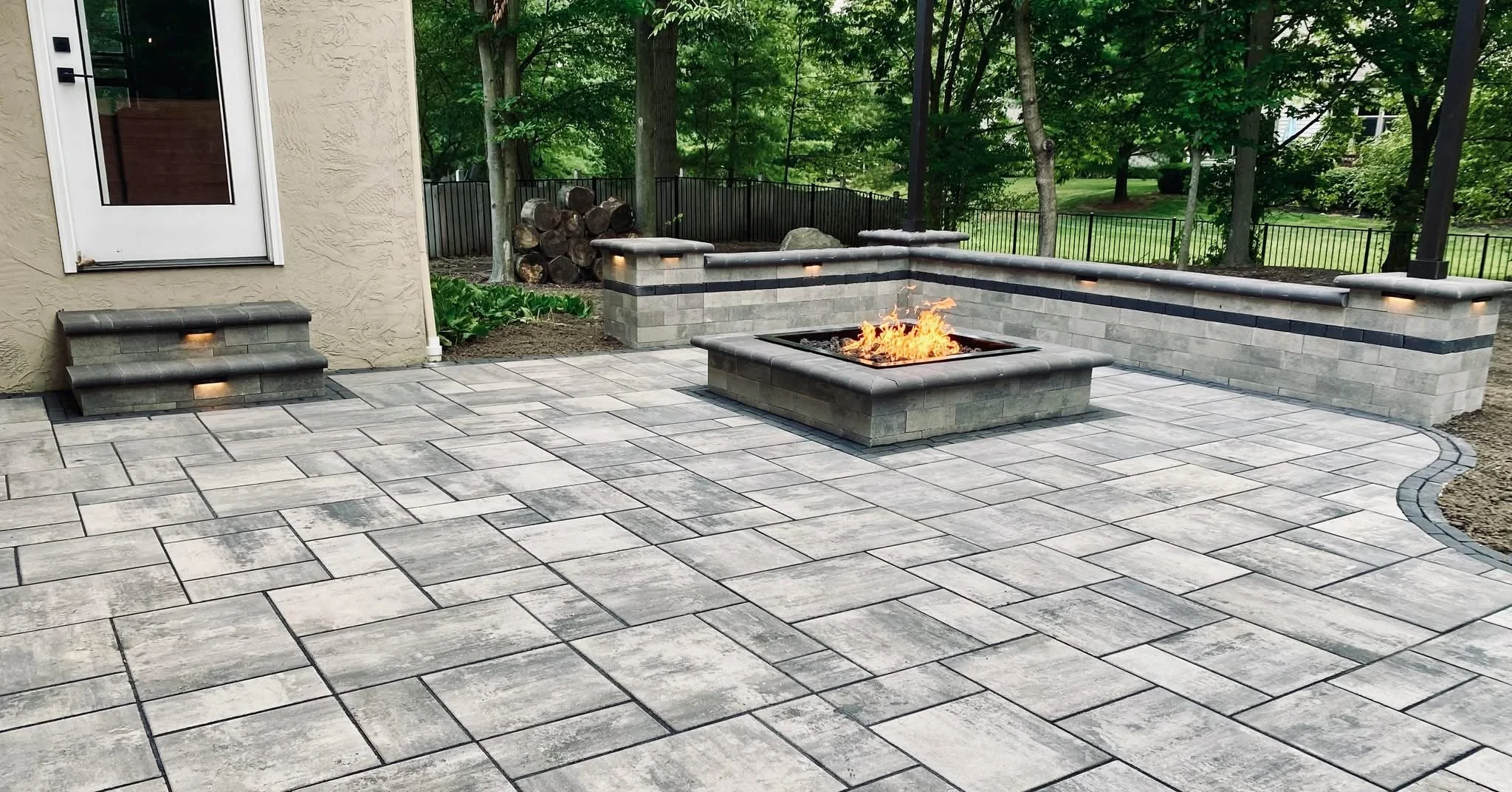 Backyard patio with patterned concrete pavers, a fire pit, and stone seating wall, surrounded by green trees and a black metal fence.