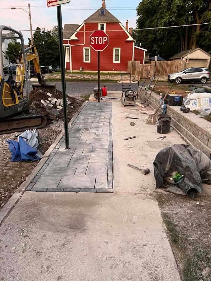 A sidewalk under construction with new paving stones installed on the left side, a stop sign at the street intersection, and construction equipment and materials scattered around. A red house and parked cars are visible in the background.