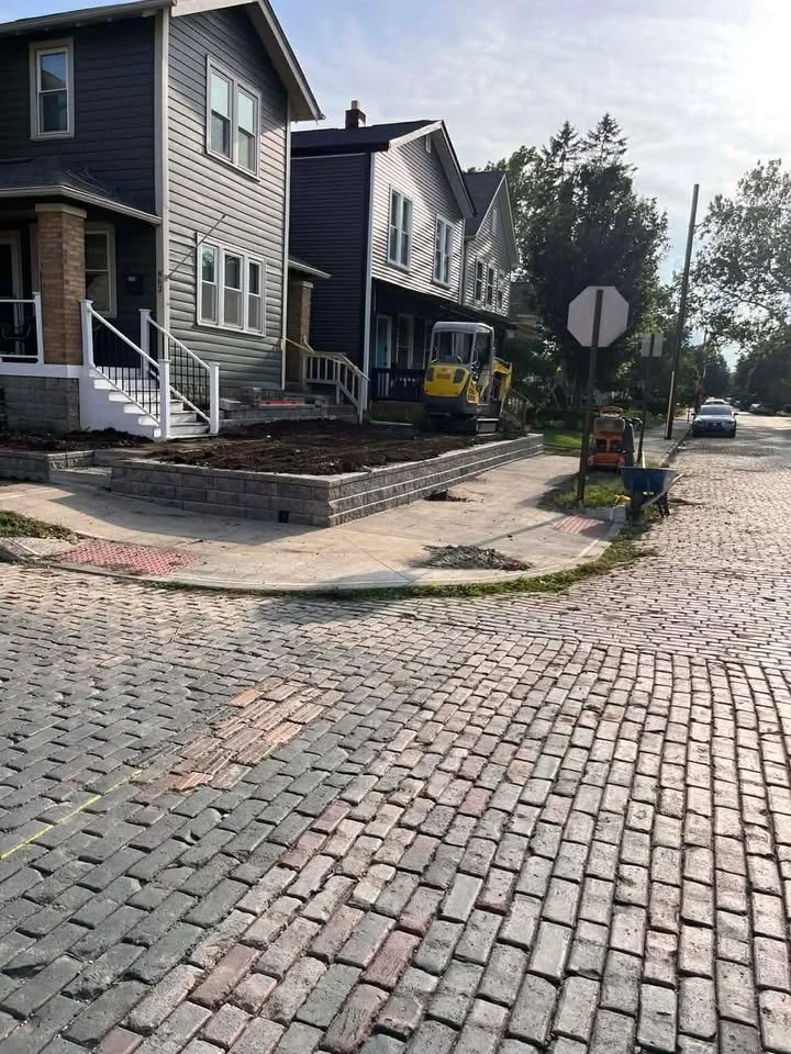 Newly paved and landscaped front yard of a residential house under construction, with construction equipment and tools visible, on a cobblestone street.