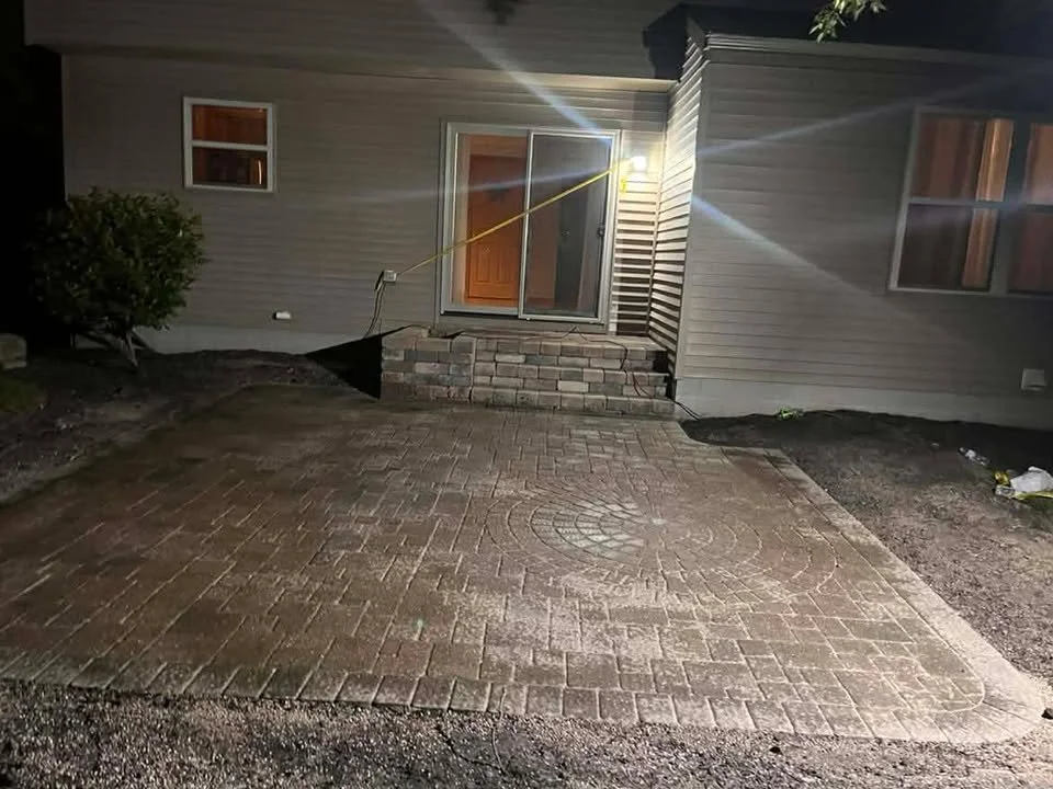 Nighttime view of a newly paved brick patio in front of sliding glass doors leading into a house, with a light on the exterior wall illuminating the area.