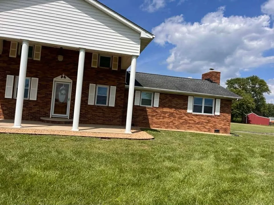 A brick house with white shutters and a porch with white columns, surrounded by a green lawn under a partly cloudy sky.