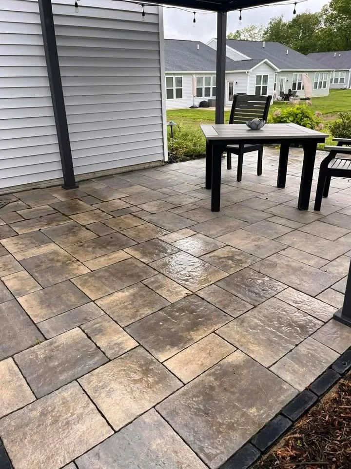 Backyard patio with stone tiles, outdoor table with a bowl, and chairs, with neighboring houses and greenery in the background.