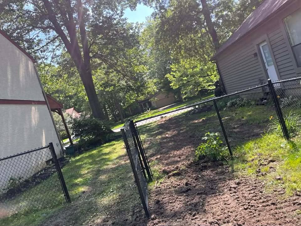 Yard with a partially assembled black chain-link fence, a large tree, and neighboring houses in a suburban neighborhood during daytime.